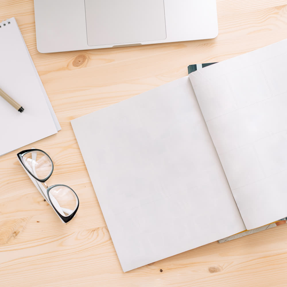 An open book, a notepad and pen, reading glasses, and a laptop laid out on a light wood desk